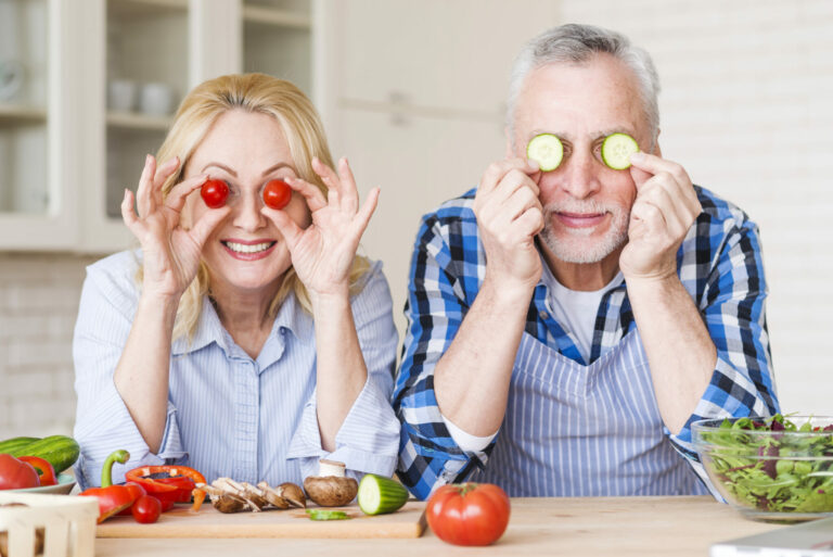 smiling-senior-couple-holding-cherry-tomatoes-cucumber-slices-front-their-eyes
